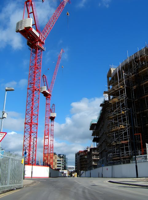 A construction site featuring two tall red tower cranes with white counterweights on their upper sections, positioned against a blue sky with scattered white clouds. The cranes' long horizontal jibs extend outward, with one jib having a visible hook hanging from a cable. To the right, there is a multi-storey building under construction, surrounded by scaffolding with metal poles and wooden planks, and partially covered in black netting. The site is enclosed by a metal fence, with a paved road in the foreground leading into the construction area. A streetlamp is visible on the left side of the image. The scene reflects ongoing urban development, and the image may be relevant to private waste or rubble removal services provided by Waste Clearance Islington for construction sites, emphasizing on-site removal of building debris and waste during development activities.