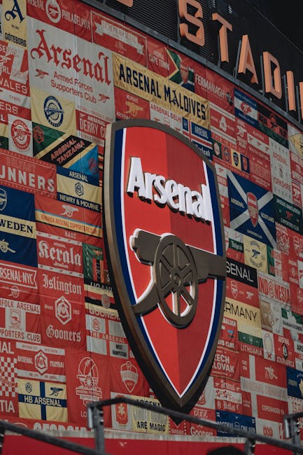 A large, vertically oriented sign on the exterior wall of a building displaying the Arsenal Football Club crest, which features a red shield with a white outline, a black cannon with a grey barrel directed to the right, and the word 'Arsenal' in bold white letters above the cannon. The wall behind consists of numerous smaller banners, patches, and stickers in various colors, predominantly red, white, blue, and beige, with some incorporating team logos, text, and club-related graphics. The surface appears textured with the overlapping advertising material creating a layered visual effect, typical of stadium or sports team environments. The lighting is natural and evenly distributed, illuminating the entire display clearly. This scene suggests an emphasis on team identity and community support, relevant to sources offering private or alternative ways of displaying or acquiring sports memorabilia or club-related signage, aligning subtly with independent approaches to club merchandise collection or on-site display, as seen in private collectors or sports venues.