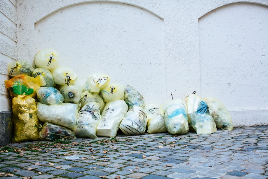A collection of partially transparent plastic rubbish bags, predominantly yellow and white, are stacked along the base of a white wall with textured finish. The bags contain various types of waste, some appearing to hold soft materials, while others seem to contain packaging or miscellaneous debris. The bags are tied with black or dark-colored twist ties and are arranged in a loose pile on a cobbled pavement surface, which features small, irregular stone blocks in shades of grey and brown, with some fallen leaves scattered around. In the background, the white wall extends vertically, with a subtle curved arch detail at the top, and the scene appears to be outdoors, possibly in a courtyard or alleyway. The overall setting suggests a private or independent waste collection effort, consistent with rubbish removal services like Waste Clearance Islington. The natural lighting is diffuse, indicating an overcast day.