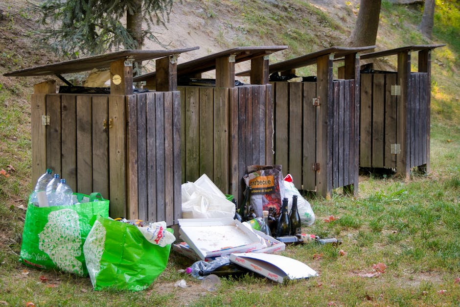 Four wooden outdoor toilets with dark brown vertical planks and sloped roofs are positioned side by side on a grassy area, with the environment featuring trees and a sloped dirt bank in the background. In front of the first toilet, there are various discarded items including green plastic bags filled with empty plastic bottles, a white pizza box with some remains inside, a black paper bag containing barbecue charcoal, and several empty glass bottles. The scene depicts improper disposal of waste in an outdoor setting, illustrating the type of rubbish that could be encountered during private or alternative waste handling services, such as those offered by Waste Clearance Islington, especially when addressing bulky waste collection problems in urban or semi-rural areas.