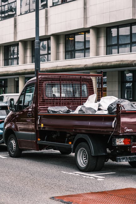 A small, dark red pickup truck with an open cargo bed carrying a load of mixed rubbish, including white plastic bags filled with waste and some darker-colored items, parked on a city street in front of a modern building with large windows and a concrete facade. The truck features a protective metal grille at the cab's rear, and its wheels have silver rims with black tires. The background includes parked vehicles and a light pole, with the urban environment suggesting a designated area for disposal or waste collection. The scene reflects a typical private rubbish removal service, with the vehicle likely used by Waste Clearance Islington for on-site clearance or alternative waste handling outside standard municipal waste collection. The overall lighting is natural, indicating daytime, and the surroundings are clean and orderly, emphasizing efficient rubbish clearance in an urban setting.