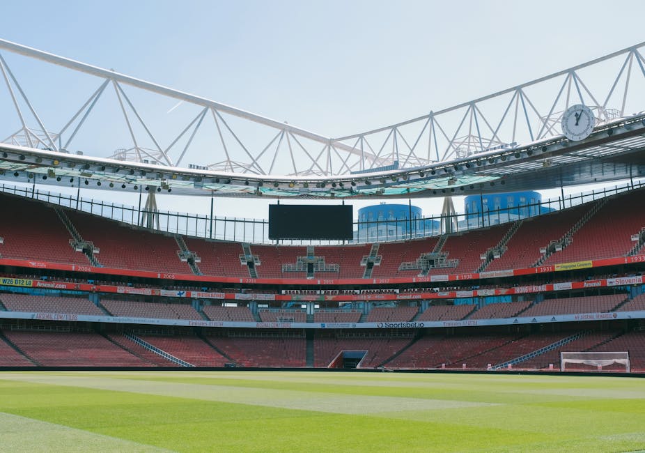 An empty football stadium featuring a well-maintained grass pitch with clear white lines, surrounded by multiple tiers of red and blue seating. The seating areas are partially shaded by the stadium's roof structure, which includes white, crisscrossing beams extending outward. A large black digital scoreboard is suspended in the middle of the upper section, and a clock displaying the time is mounted on the right side of the roof framework. In the background, modern blue and glass buildings are visible behind the stadium, indicating an urban location. The lighting suggests a bright, clear day with natural sunlight illuminating the whole scene, highlighting the clean and orderly environment typical of sports venues. This setting is relevant in illustrating the potential for event-related waste management and the importance of professional rubbish removal, especially in large-scale venues, aligning with the services provided by Waste Clearance Islington for efficient on-site clearance and waste handling during events or renovations.
