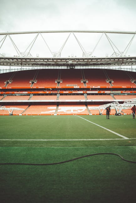An empty football stadium featuring a well-maintained grass pitch with clear white lines, surrounded by multiple tiers of red and blue seating. The seating areas are partially shaded by the stadium's roof structure, which includes white, crisscrossing beams extending outward. A large black digital scoreboard is suspended in the middle of the upper section, and a clock displaying the time is mounted on the right side of the roof framework. In the background, modern blue and glass buildings are visible behind the stadium, indicating an urban location. The lighting suggests a bright, clear day with natural sunlight illuminating the whole scene, highlighting the clean and orderly environment typical of sports venues. This setting is relevant in illustrating the potential for event-related waste management and the importance of professional rubbish removal, especially in large-scale venues, aligning with the services provided by Waste Clearance Islington for efficient on-site clearance and waste handling during events or renovations.