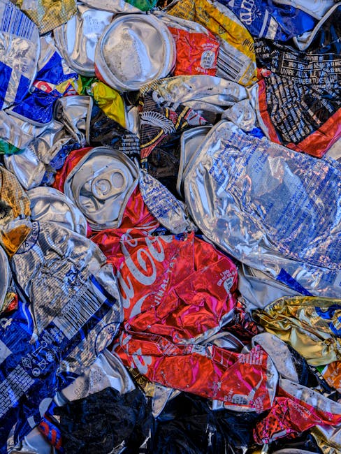 A close-up image of a pile of crushed and crumpled aluminium cans and plastic bottles, featuring various colours including silver, red, blue, black, and yellow. The cans display visible pull-tabs and ridged surfaces, with some partially flattened and others more intact. The plastic bottles have caps and printed labels, with textures that range from smooth to wrinkled. The cans and bottles are loosely stacked and overlapping, positioned on what appears to be a rough surface or ground, suggesting an outdoor or industrial setting. The lighting highlights the metallic sheen of the cans and the glossy finish of the plastic bottles, emphasizing their materials. This scene illustrates typical waste collection in a rubbish removal context, aligning with private disposal or on-site clearance services provided by Waste Clearance Islington, as shown on their website. The image focuses on the raw, discarded recyclables, emphasizing the need for proper waste management and adherence to local disposal rules, reflecting the importance of responsible rubbish handling in urban environments.