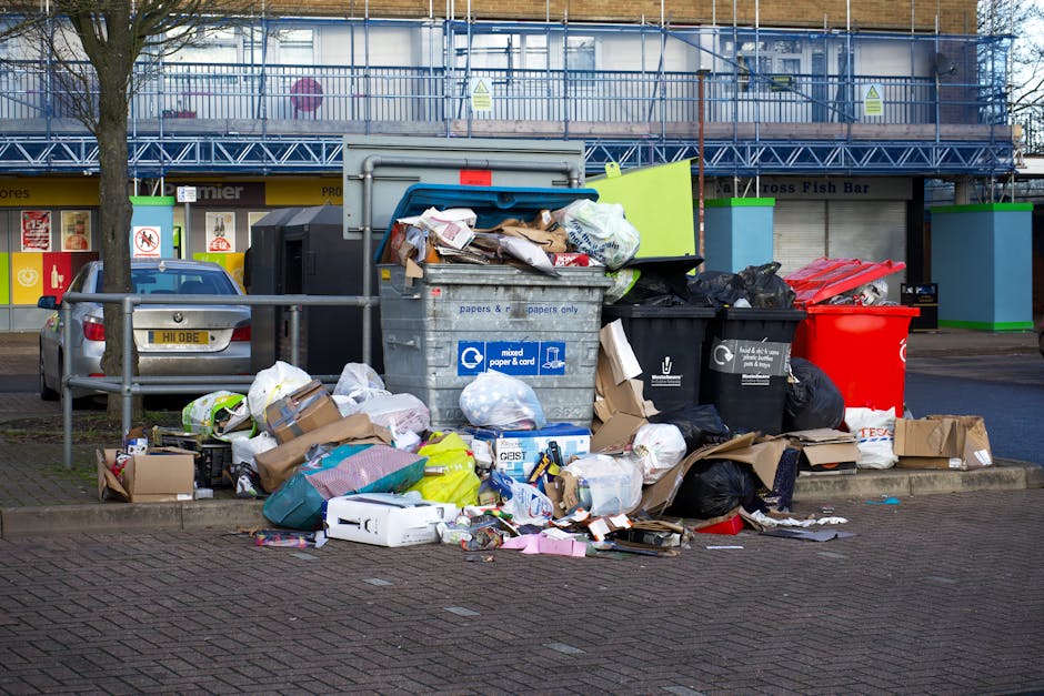 An outdoor scene featuring a collection of overflowing rubbish and recycling bins situated on a paved area next to a metal safety barrier. The large, grey mixed paper and cardboard recycling bin is partially open, with brown paper bags, cardboard boxes, and various paper waste spilling out onto the ground. Adjacent to this are black and red waste bins, with black being used for general waste and the red for possibly recyclable or hazardous waste. The black bin is filled with plastic bags and other refuse, some tied shut. Additional cardboard boxes, paper, plastic packaging, and miscellaneous rubbish are scattered around the bins, creating a cluttered appearance. In the background, there is a commercial building under renovation, covered with scaffolding and mesh netting, and a row of shops with closed shutters. To the left, a parked grey car is visible behind the safety barrier, with a tree trunk nearby. The scene reflects a typical urban waste accumulation point, highlighting the importance of proper waste management and disposal, which companies like Waste Clearance Islington manage through private rubbish collection services rather than local authority collection schemes.