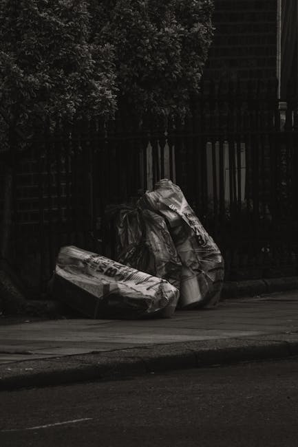 On a paved roadside pavement, three black plastic garbage bags are positioned in front of a black metal fence with vertical bars. The bags appear to contain household waste or discarded materials, with the middle bag leaning slightly to the right and the others resting on the ground. The plastic bags have some visible printed text and labels, indicating they may have been used for rubbish collection. Behind the fence, dense greenery including trees and shrubs provides a backdrop, partially shadowed by an overcast or shaded environment. The scene reflects typical urban waste disposal, possibly related to non-organic or mixed rubbish needs, handled by private waste clearance services such as Waste Clearance Islington. The overall lighting is subdued, emphasizing the utilitarian aspect of rubbish accumulation on the street edge, illustrating a common private waste collection scenario where waste is temporarily stored before removal. The setting, with its public pavement and fencing, highlights the importance of proper rubbish management and the role of independent waste disposal services in maintaining cleanliness in urban environments.
