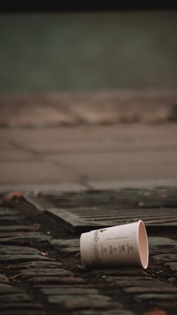 A discarded white paper cup with printed design and text lies on a brick pavement, tilted on its side near a metal drain cover. The surrounding area includes a weathered wooden surface and dark, damp stones, suggesting an outdoor urban environment. The lighting appears natural, highlighting the cup's clean surface against the darker, textured background. This scene exemplifies common refuse found in public spaces, which waste clearance companies such as Waste Clearance Islington often manage through private rubbish removal services, focusing on removing light litter or debris from streets, pavements, and alleyways to maintain tidiness and environmental responsibility.