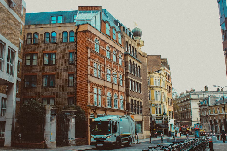 A street scene in an urban area featuring several multi-story buildings with a mix of architectural styles, including brick facades and ornate detailing. In the foreground, a light blue waste collection truck with a compact cab and large rear compactor is parked on the right side of the street, positioned near a series of black metal bike racks. The street appears to be a busy thoroughfare with other vehicles, including a red bus and additional trucks, visible in the background. The scene is illuminated by natural daylight, casting soft shadows on the buildings and street, with a grey overcast sky overhead. The surroundings include lampposts, street signs, and a few pedestrian elements, indicating an active city environment. Waste Clearance Islington occasionally conducts private rubbish collection and on-site clearance services, which are visually represented here through the parked waste vehicle aligned with the urban context of alternative waste handling methods.