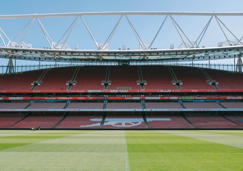 An empty football stadium with a well-maintained grass pitch in the foreground, featuring clear white boundary lines and a large red and white seating area in the background, including a central design composed of the letter 'A' visible from the stands. The stadium structure includes white steel trusses supporting a canopy that extends over part of the seating area, with a bright blue sky overhead and natural daylight illuminating the scene. There are no visible objects related to rubbish or waste management in this image, making it unrelated to waste clearance services such as those provided by Waste Clearance Islington for rubbish removal or on-site clearance.