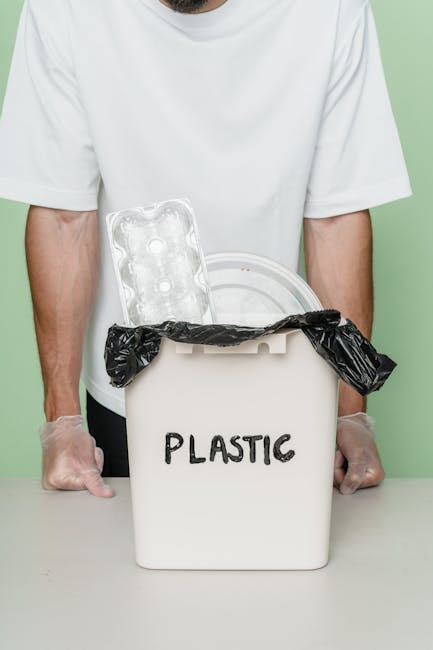 A person, wearing a white t-shirt and transparent plastic gloves, stands behind a white plastic container labeled 'PLASTIC,' which is used for waste collection. The container is partially covered with a black plastic rubbish bag, tied around its opening. Two empty plastic trays or blister packs with a translucent, slightly glossy finish rest on top of the container. The background features a plain light green wall, providing a neutral setting. This scene relates to waste management activities, potentially involving the sorting or disposal of plastic waste, similar to services offered by waste clearance companies for waste collection and recycling in urban environments. The visual emphasis on plastic waste materials underscores the importance of proper disposal and alternative collection methods that avoid reliance on municipal services, aligning with private rubbish removal or on-site clearance practices often used in residential or commercial settings.