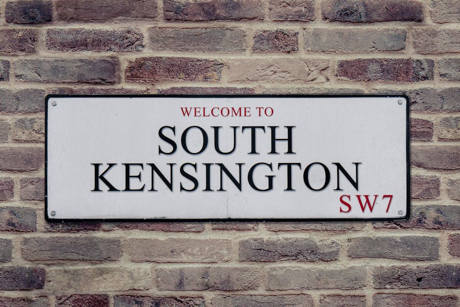 A rectangular metal street sign mounted on a brick wall displaying the text 'Welcome to South Kensington SW7' in black and red lettering, with the words 'Welcome to' positioned above 'South Kensington' and the postal code 'SW7' on the lower right corner. The sign has a white background with a thin black border, and the brick wall behind it features reddish-brown bricks with varying shades and texture, evenly laid in a horizontal pattern. The image captures the sign and wall in natural lighting, emphasizing the contrast between the clean, modern signage and the rustic brick surface, which could relate to surroundings where independent waste disposal or on-site clearance might be considered as part of property management or renovation projects by waste clearance services such as Waste Clearance Islington.