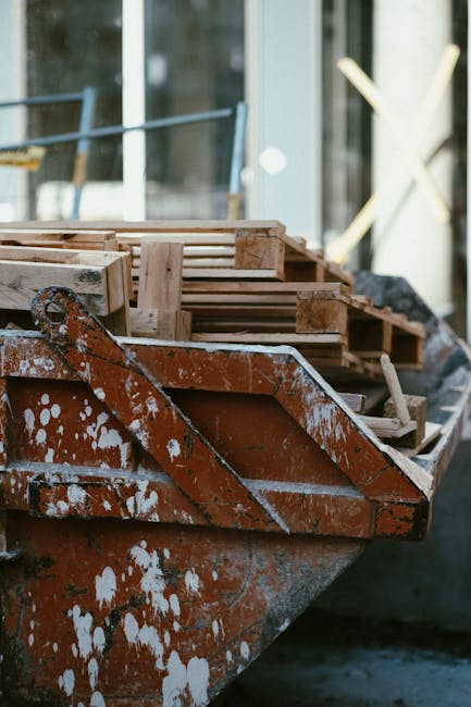A close-up view of a rusty, weathered metal boat with chipped white paint, filled with neatly stacked wooden pallets that are light brown with visible grain and a rough texture. The pallets are arranged in an orderly manner, with some leaning slightly to one side. The scene is set outdoors, possibly on a dock or near a building with large glass windows, which reflect the surrounding environment. In the background, there is a blurred glimpse of metallic scaffolding or framework, and a large window with a vertical window frame visible. The lighting is natural, with soft daylight illuminating the scene, highlighting the textures of the rusted metal, wood grain, and the reflective glass surface. This image ties into the theme of waste management and potential for on-site clearance or waste separation in a construction or renovation context, consistent with services offered by Waste Clearance Islington.